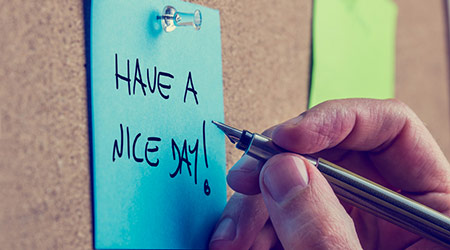 Closeup of male hand writing Have a nice day message on a blue post it paper pinned to a cork bulletin board, toned retro.