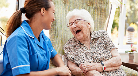 Senior Woman Sitting In Chair And Talking With Nurse In Retirement Home