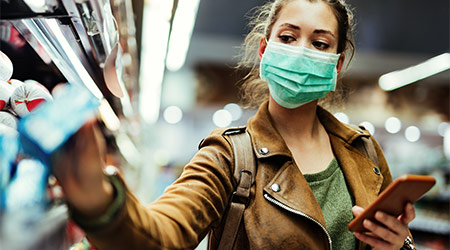 Young woman with face mask using mobile phone and buying groceries in the supermarket during virus pandemic.