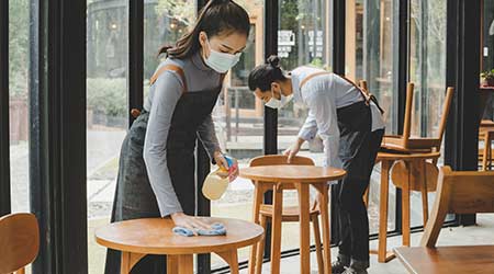 asian waitress staff wearing protection face mask in apron cleaning table with disinfectant spray for protect infection coronavirus (covid-19) in cafe coffee shop restaurant, hotel. new normal concept