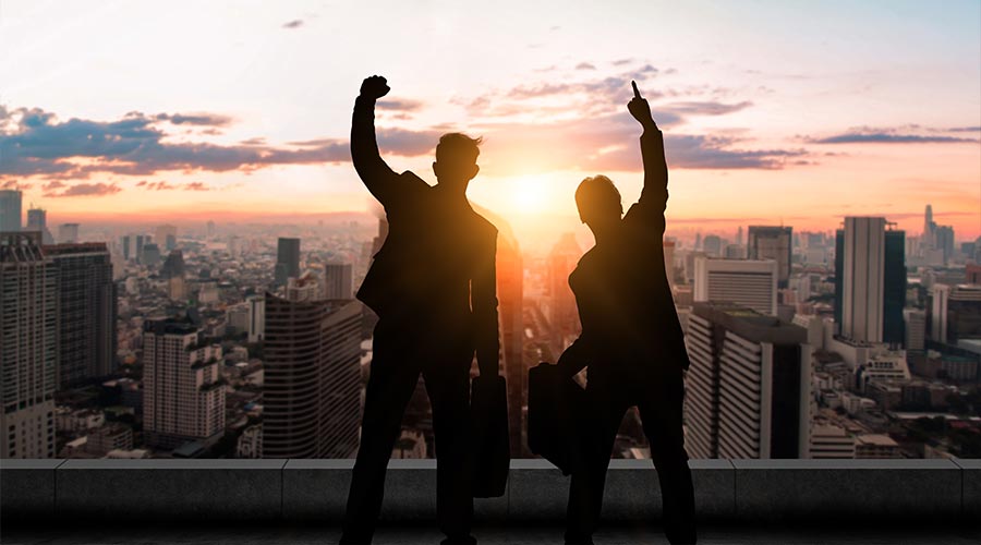 Two silhouetted businessmen celebrate their success on a rooftop with a city skyline and sunset in the background