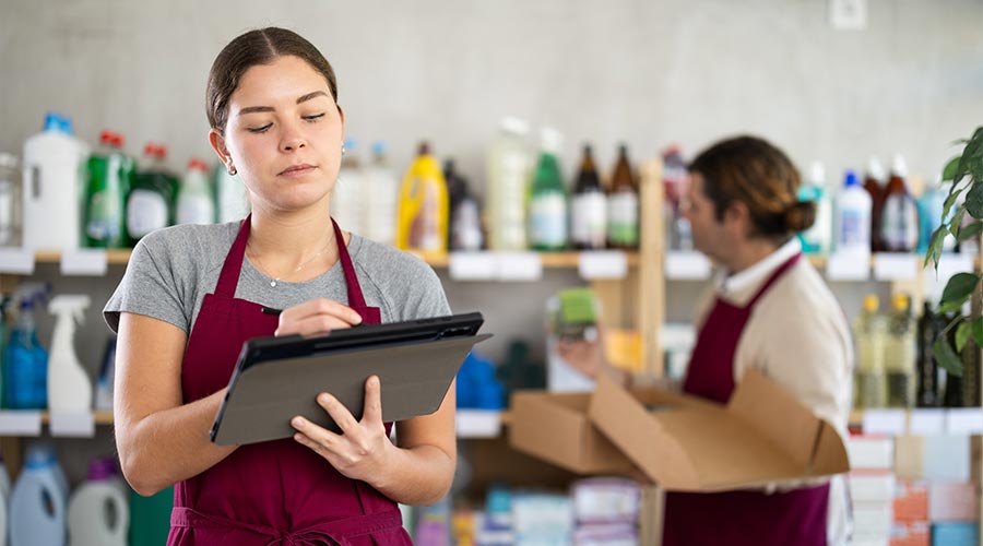 Young female store supervisor in burgundy apron conducting inventory checks on cleaning and hygiene products using tablet, with male staff member working in background