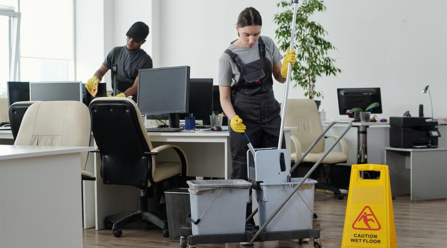 Young cleaning lady sqeezing mop in bucket against male colleague while going to wash floor in large modern openspace office